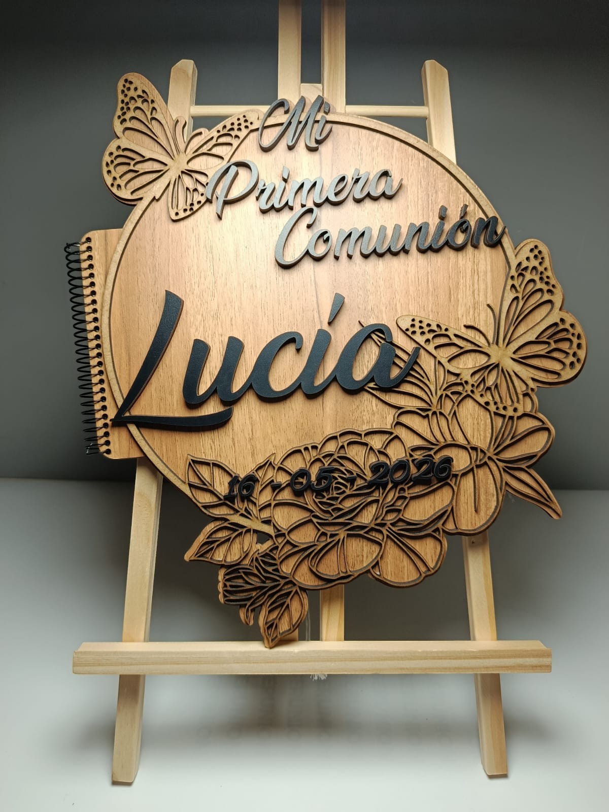 Álbum de comunión de madera con detalles 3D de mariposas y flores. Incluye el nombre 'Lucía' y la fecha 10-05-2020. Izmi Láser Regalos Personalizados.